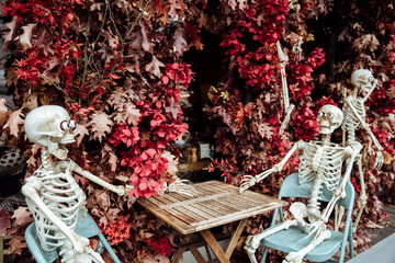 Halloween decoration near cafe or restaurant, modern skeletons sitting, talking, using mobile phones, having fun like people outdoor.