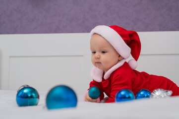 Obraz premium Baby boy dressed as Santa is playing with Christmas decorations on the bed.