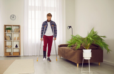 Man with broken leg, sprained ankle or foot injury walking with crutches at home. Young guy wearing ankle support brace or fracture fixator with adjustable straps walking on his own in living room - obrazy, fototapety, plakaty