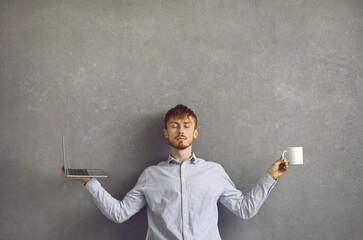 Reduce stress at work: Portrait of calm male corporate employee or entrepreneur meditating sitting in yoga pose arms outspread and holding laptop computer and coffee in hands against grey studio wall