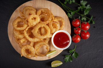 Homemade baked onion rings fries with mayonnaise, Tomato sauce and rosemary on wooden board. fast food products : onion rings on cutting board, on black stone background, unhealthy food.