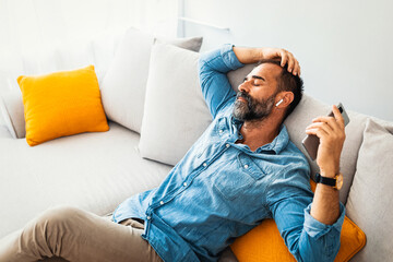 Happy relaxed man with a blissful smile sitting listening to music on his mobile phone using an earbud as he sits on a sofa at home. Man relaxing holding using phone enjoy favorite songs relaxing