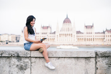 Tourist reading book while sitting on retaining wall with government building in background, Budapest, Hungary