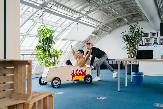 Colleague pushing cheerful businessman sitting in toy car at creative office