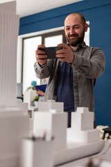 Naklejka na ściany i meble Professional architect holding smartphone smiling in videocall conference presenting final maquette to client. Bearded architectural engineer photographing table with white foam residential model.
