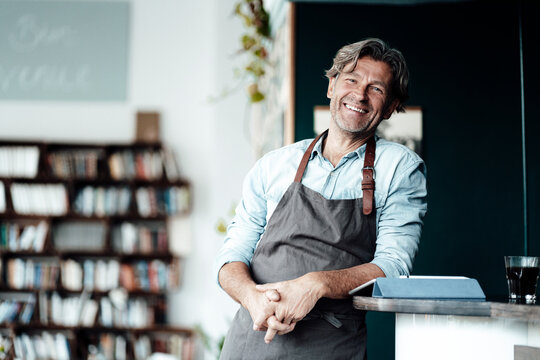 Cheerful male cafe owner wearing apron leaning on table