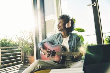 Smiling woman listening music through headphones while playing guitar at home