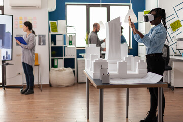 Architect standing in modern architectural office using virtual reality goggles to view 3d plan of white foam maquette. Engineer working with vr headest next to table with model of urban project.