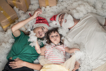 Obraz premium Christmas photo of happy Family. New Year Portrait of dad, mom and daughter wearing Christmas red hats, smiling and laying in white artificial snow. 