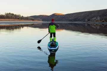 A man is rafting on the river on a supboard in the late autumn in the evening, looking into the distance at the mountains. SUP