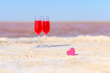 Small pink heart on the background of glasses with rose wine and pink lake. Romantic getaway concept. Selective focus on the heart.