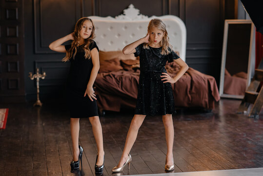 Two beautiful little girls in black dresses on mother's heels are posing in the studio against the background of the bed. Style and fashion concept. Children put on mature shoes. Photo in motion.