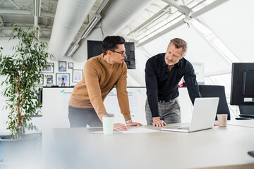 Male professionals with laptop discussing while leaning on desk in office