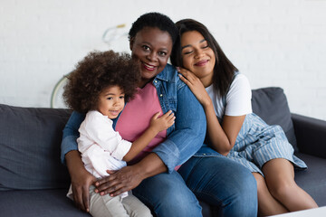 mature african american woman smiling at camera while embracing on sofa with young daughter and toddler child