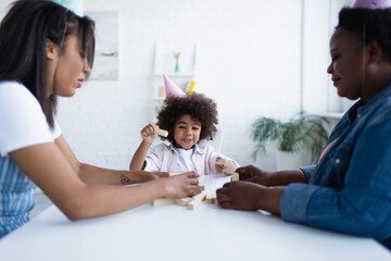 positive african american girl in party cap playing wood blocks game with blurred mom and granny