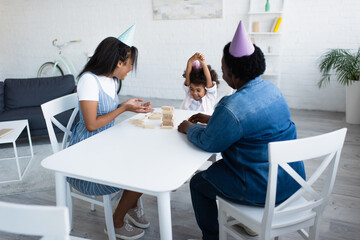 african american woman in party cap pointing at wooden tower on table near happy daughter