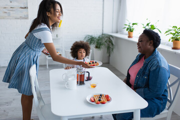 smiling african american woman holding plate with waffle and berries near mom and daughter