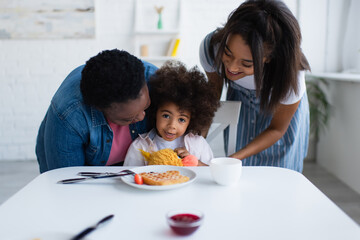 happy african american women embracing kid sitting with soft toy near waffle and bowl with jam