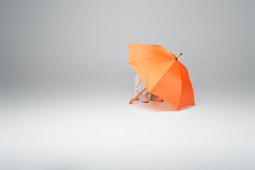 kid in white clothes hiding under orange umbrella on grey background with copy space