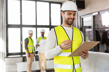 architecture, construction business and building concept - happy smiling male architect in helmet and safety west with clipboard working at office