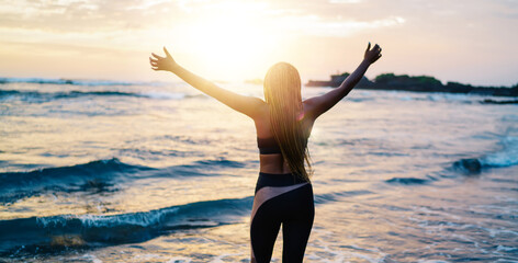 Back view of satisfied female runner in sportive tracksuit raising hands during workout break at seashore coastline, carefree woman with casual figure enjoying slimming goals and own triumph