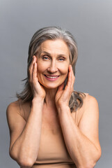 Happy carefree mature lady stands in studio, isolated on grey. Charming beautiful active and fit mature gray-haired woman holding face with hands, looks at the camera, smiles toothy. Face aging care