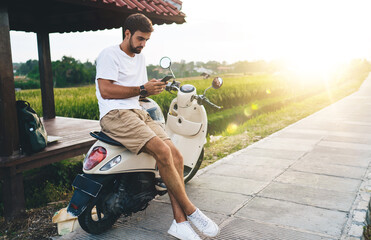Focused tourist chatting on smartphone on scooter in countryside