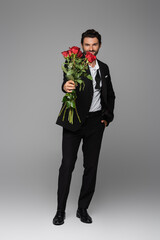 full length of happy man in suit holding red roses while standing with hand in pocket on grey