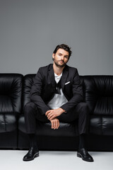 bearded man in suit sitting on black leather sofa on grey