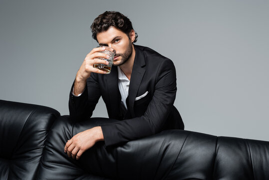 bearded man in suit standing near black sofa and holding glass of whiskey isolated on grey