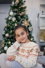 Adorable dark haired little girl 9 years old in pajamas near the Christmas tree. New Year's lights. Winter holidays.