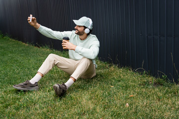 positive man in wireless headphones taking selfie while sitting on green grass with paper cup