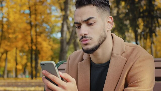 Portrait young serious guy use app hispanic man looking at phone screen browses news on telephone concentrated male sits on bench in autumn park read using device businessman holds smartphone outdoors