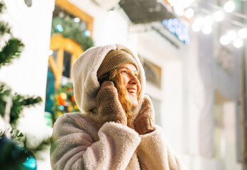 Portrait of young happy woman with curly hair in fur jacket having fun in the winter street decorated with lights