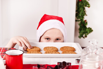 Kid boy in Santa cap taking try to steal cookie from table.