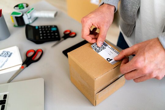 Male entrepreneur sticking QR code on cardboard package at desk