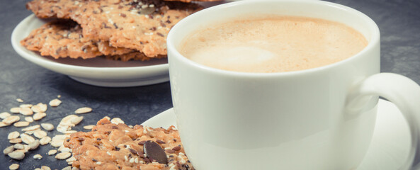 Cup of coffee with milk and fresh oatmeal cookies with honey and healthy seeds on white plate. Delicious crunchy dessert