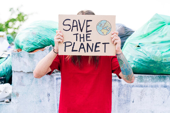 Hippie man covering face with save the planet cardboard near garbage dump