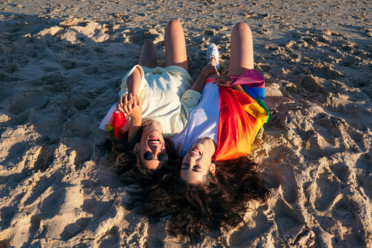 Happy female friends holding hands while lying on rainbow flag at beach