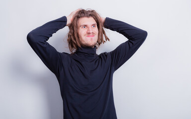 Surprised funny man holding his head, puffed out cheeks and compressed lips. Man in dark gray turtleneck and short dreadlocks on head hold hands to his head on a gray background copy space