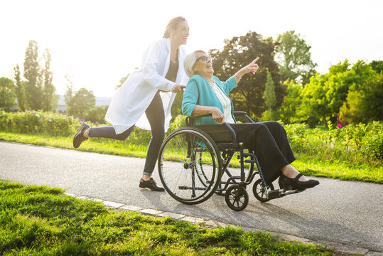 Cheerful senior woman pointing while caretaker wheeling wheelchair in park