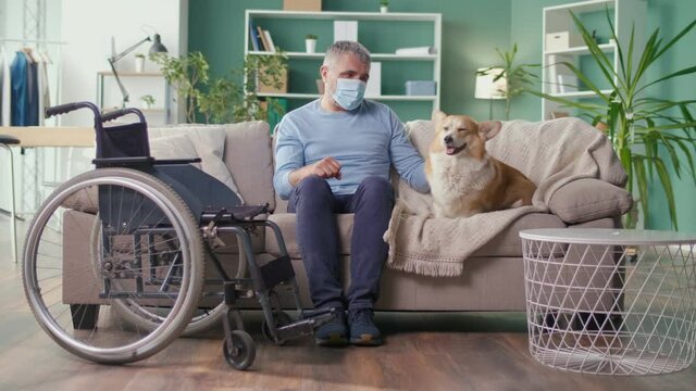 A Sad Elderly Man with Disabilities in a Medical Mask Stroking His Dog While Sitting on the Couch. Canistherapy, Best Friend, Pet, Consolation, Help. Pandemic, Social Distancing and Self-isolation.