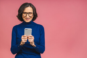 Naklejka premium Happy mature senior woman holding smartphone using mobile online apps, smiling old middle aged lady texting sms message chatting on phone looking at cellphone isolated over pink background.