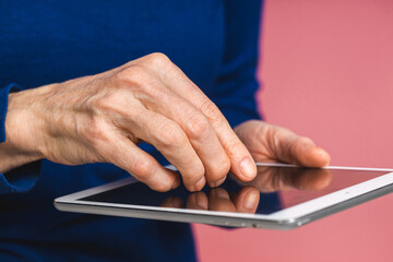 Close-up photo of smiling beautiful senior aged mature woman using tablet computer, isolated over pink background. Copy space for text.