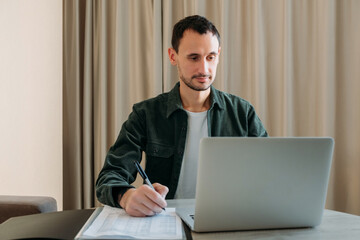 Portrait of a contented young man using a laptop at a table in office or at home. Work at home concept. Filling out documents, checking through a computer.