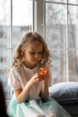 A girl cleans a tangerine sitting by the window