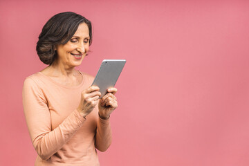 Portrait of smiling beautiful senior aged mature woman using tablet computer, isolated over pink background. Copy space for text.