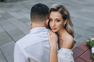 Stylish groom and cute blonde bride in a white dress are hugging while sitting on a bench on the street in the city. Wedding photography of the newlyweds.