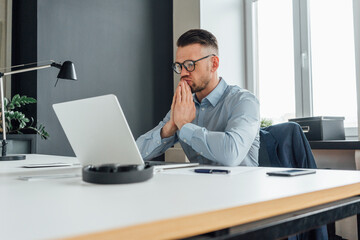 Thoughtful businessman with hands clasped looking at laptop in office