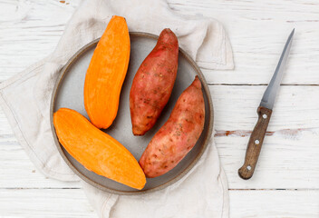 Raw organic whole and sliced sweet potatoes in a gray plate on a white wooden background. Fresh Root vegetables. Selective focus, top view and copy space